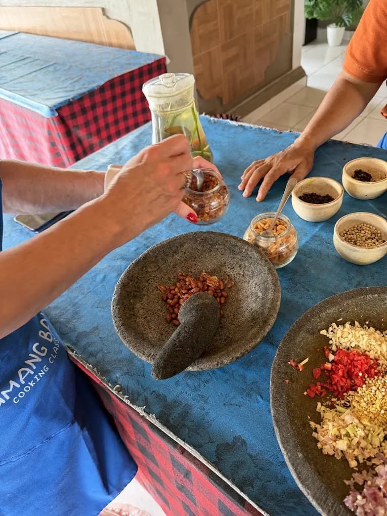 Hands grinding base genep in a stone mortar at the Petulu spice table, from the Petulu class where sambal embe is cooked