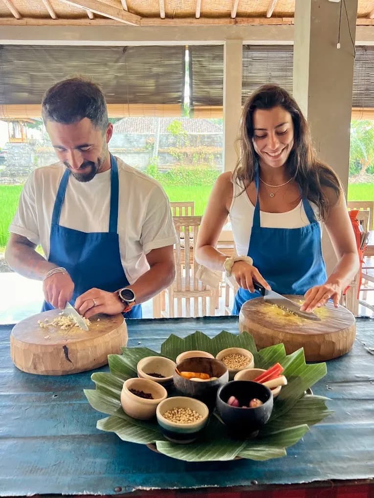 Two guests in blue aprons chopping garlic on wooden blocks beside a banana-leaf tray of spices, rice field behind, in Petulu
