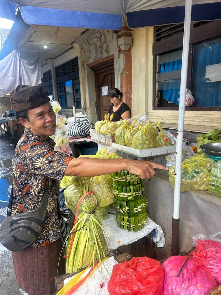 A morning market stall piled with chilies, shallots, and garlic, with a guide in a blue apron, from the Petulu class where klepon is cooked